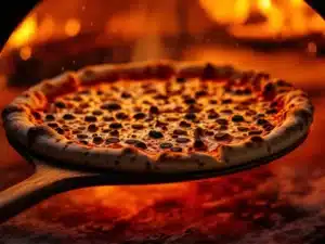 Napolitaanse pizza met geplekte korst wordt op houten schep in vurige pizzaoven geschoven met zichtbare vlammen