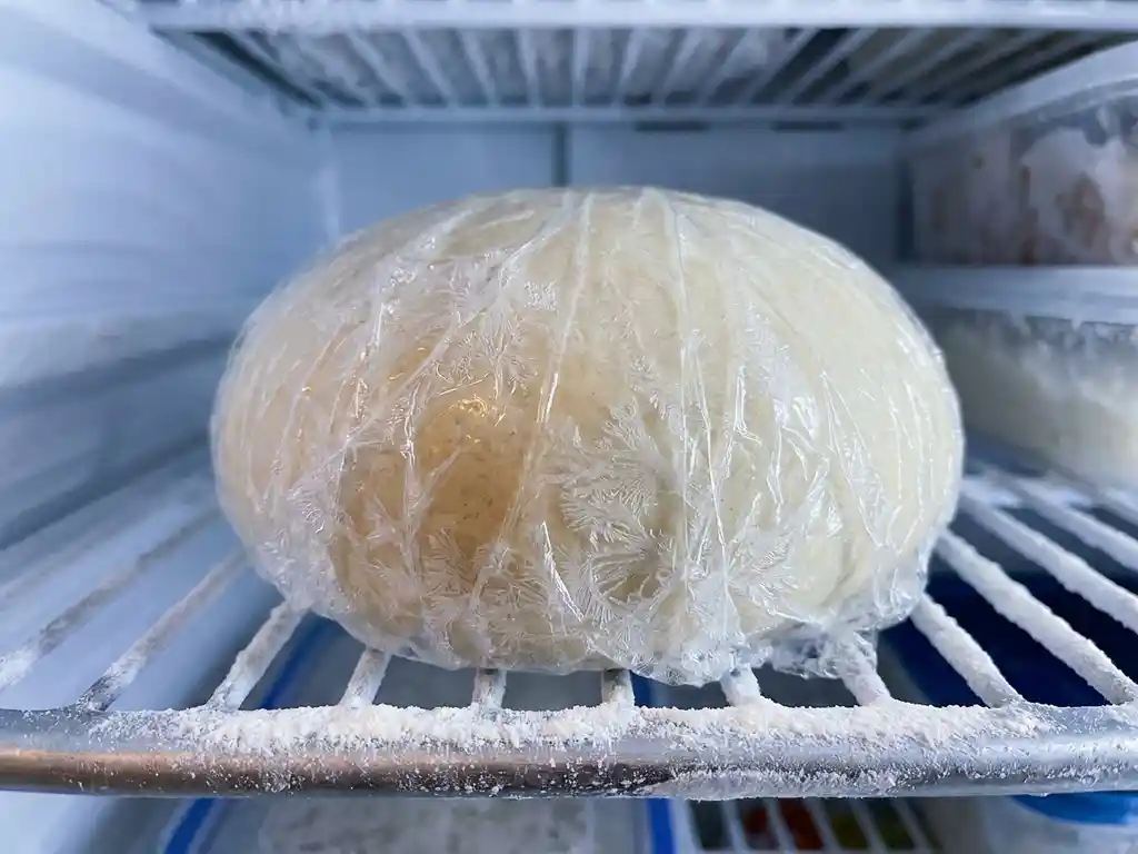 Glutenvrij pizzadeeg in plasticfolie op een bevroren vriezerplank met ijskristallen en een laagje bloem.